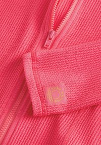 Giacca a zip di un brillante colore rosa con un motivo testurizzato. Presenta una cerniera rosa e un'etichetta con il logo "SURF NATION" in giallo. Tessuto morbido.