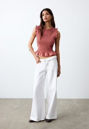 Donna che indossa un top rosa senza maniche con volant e pantaloni bianchi a gamba larga, in piedi contro una parete bianca semplice e un pavimento grigio.