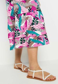 Blumendruckkleid in Pink-, Teal- und Grautönen; mit geschichteten Rüschen am Saum. Weiße Sandalen mit Riemchen und flacher Sohle.