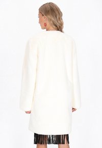 Kvinde iført en lang hvid faux fur-frakke over en sort frynseskørt, vendt væk med bølget blond hår og synlige røde hoop øreringe.