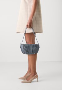 Denim handtas met geplooid ontwerp, versierd met zilveren studs, gedragen door een persoon in een geruite jurk en naakte hakken, voor een neutrale achtergrond.