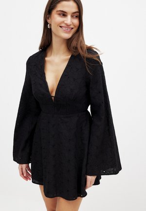 Nainen pukeutuneena mustaan pitkähihainen kukkakuvioiseen pitsiminimekkoon, jossa on syvä V-pääntie, hymyilee ja katsoo sivulle valkoista taustaa vasten.