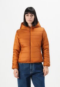 Oranje pufferjack met capuchon, ritssluiting en gequilte afwerking. Voorzien van elastische manchetten en een licht getailleerd model. Gekleed met blauwe jeans.