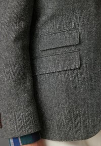 Grau karierter Blazer mit zwei horizontalen Taschen, aus strukturiertem Wollstoff. Sichtbares Bündchen zeigt ein blau-grünes karriertes Hemd darunter.