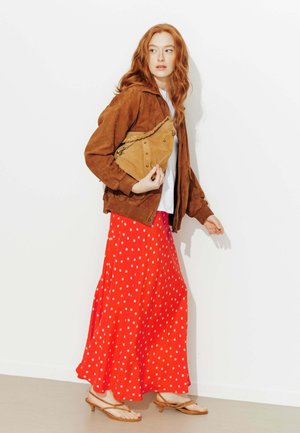 Femme aux cheveux rouges portant une veste marron, une chemise blanche, une jupe rouge à pois blancs, des sandales beiges, tenant une pochette beige, debout contre un mur blanc.