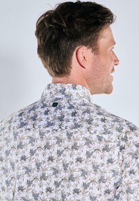 Weißes Hemd mit floralem Muster, das graue und hellbraune Blätter zeigt. Strukturierter Stoff und ein Standardkragen mit sichtbarem Etikett auf der Rückseite.