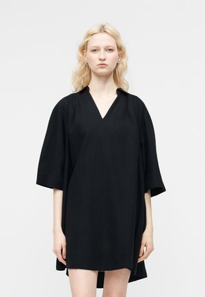 Ξανθιά γυναίκα με κυματιστά μαλλιά που φοράει ένα φαρδύ μαύρο φόρεμα με V-λαιμόκοψη και μανίκια μέχρι τον αγκώνα, σε απλό ανοιχτό φόντο.