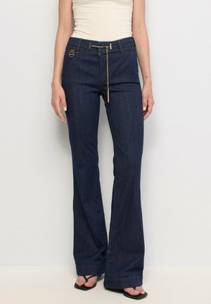 Femme portant un jean flare bleu foncé avec un détail chaîne à la taille, un top blanc sans manches et des sandales noires ouvertes, debout sur fond blanc.