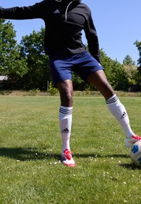 Sweat à capuche noir avec cordon blanc, shorts bleus, chaussettes blanches à trois bandes noires, chaussures de football rouges, et un ballon de football bleu sur de l'herbe verte.