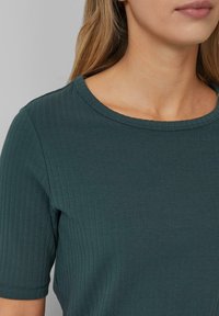 Top verde a righe con maniche corte e scollo rotondo. Il tessuto sembra morbido, con una superficie testurizzata e una leggera elasticità. Nessun dettaglio prominente visibile.