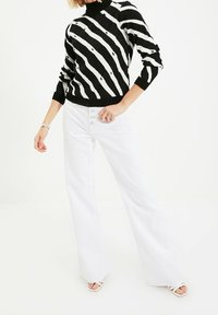 Musta-valkoraidallinen neulepusero, jossa korkea kaulus, yhdistetty valkoisiin leveälahkeisiin housuihin, joissa on edessä napit ja sileä pinta.