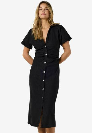 Kvinne med langt blondt hår som har på seg en svart midikjole med korte ermer og knapper foran, med rynket midje, står med hendene bak ryggen.