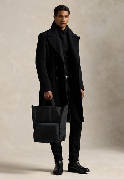 Mees mustas mantlis, kõrge kaelusega kampsunis, pükstes ja saapades, hoides käes musta esitaskuga ja logoga õlakotti, seisab beežil taustal.