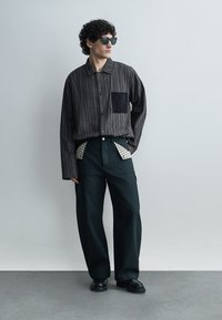 Homme aux cheveux bouclés et lunettes de soleil portant une chemise à rayures foncées avec une poche texturée et un pantalon large noir.