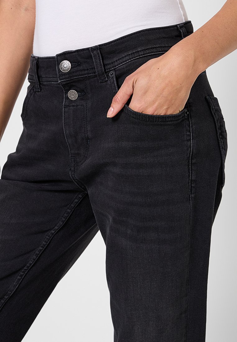 Osoba w czarnych, wysokich dżinsach z dwoma srebrnymi guzikami, z ręką częściowo w przedniej kieszeni i włożonym białym topie.