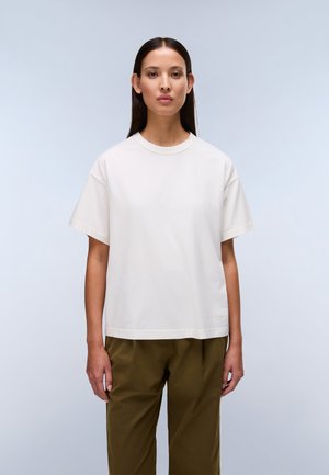 Woman with long straight hair wearing a plain white oversized T-shirt and olive green trousers, standing against a light background.