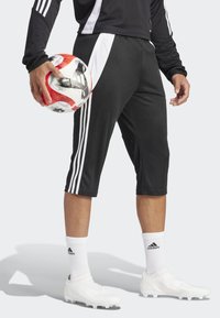 Svart idrettsshorts med hvite sidelister, iført hvite sokker og fotballsko. Spilleren holder en rød og svart fotball.