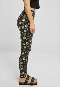 Leggings neri con motivo celestiale dorato che include soli e lune. Hanno un design aderente e sono abbinati a sandali neri e a un top corto.