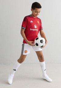 Röd Manchester United-tröja med svarta detaljer, vita shorts och svartspräckliga strumpor. Barnet håller en svart och vit fotboll.