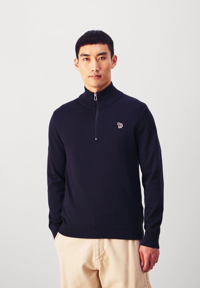 Tengerészkék pulóver magas gallérral és ezüst cipzárral, kis hímzett logóval a mellkason és bordás mandzsettákkal.