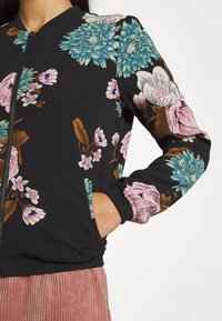 Bloemenprint bomberjack met zwarte stof en kleurrijke bloemmotieven in roze, blauw en bruin. Voorzien van een ritssluiting en zijzakken.
