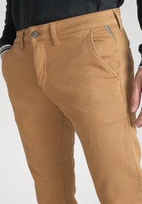 Bruna bomullsmix-byxor med knäppning, framfickor och en mjuk textur. Designen inkluderar en diskret dragkedjeficka.