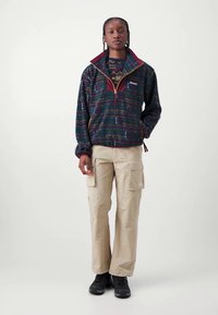 Veste à enfiler avec un motif géométrique en bleu foncé, garniture bordeaux et fermeture éclair dorée, associée à un pantalon cargo beige et des chaussures noires.
