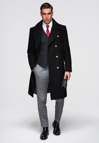 Manteau en laine noir à double boutonnage, pantalon gris sur mesure, chemise blanche, cravate rouge, gants noirs et bottines noires brillantes.