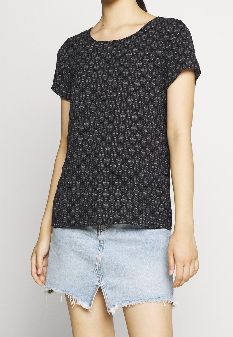 Femme portant un haut noir à manches courtes avec motifs et une jupe en jean bleu clair avec une fente à l'avant et un ourlet effiloché sur un fond uni.
