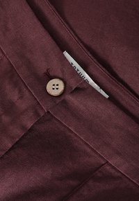 Maroon fabric with a smooth texture, featuring a brown button and visible stitching. Includes a fabric label with text.