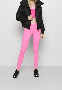 Schwarze Pufferjacke im cropped Style mit Kapuze, über einem enganliegenden pinken Sport-BH und Leggings. Die Leggings weisen strukturierte Akzente und ein elegantes Design auf.