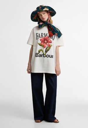 Γυναίκα φοράει άσπρη μπλούζα με φλοράλ σχέδιο, φαρδιά σκούρα τζιν, μεγάλο φαρδύ καπέλο με φλοράλ σχέδιο και δεμένη μαντήλα, καθώς και σανδάλια.