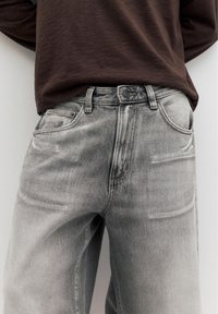 Personne portant un jean en denim gris clair et une chemise à manches longues brun foncé, les mains reposant derrière le dos.