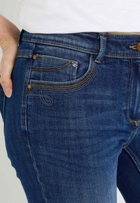 Vaqueros de mezclilla azul oscuro con un diseño ajustado, costuras detalladas en amarillo y morado, y bolsillos delanteros con remaches clásicos.