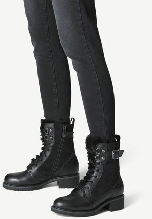 Czarne sznurowane buty sięgające do połowy łydki, z bocznym zamkiem i futrzanym wykończeniem, noszone z ciemnymi, dopasowanymi spodniami na białym tle.