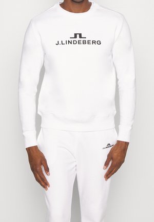 Man met witte sweatshirt en trainingsbroek met zwart J.LINDEBERG-logo op de borst en bovenbeen, staand tegen een witte achtergrond.