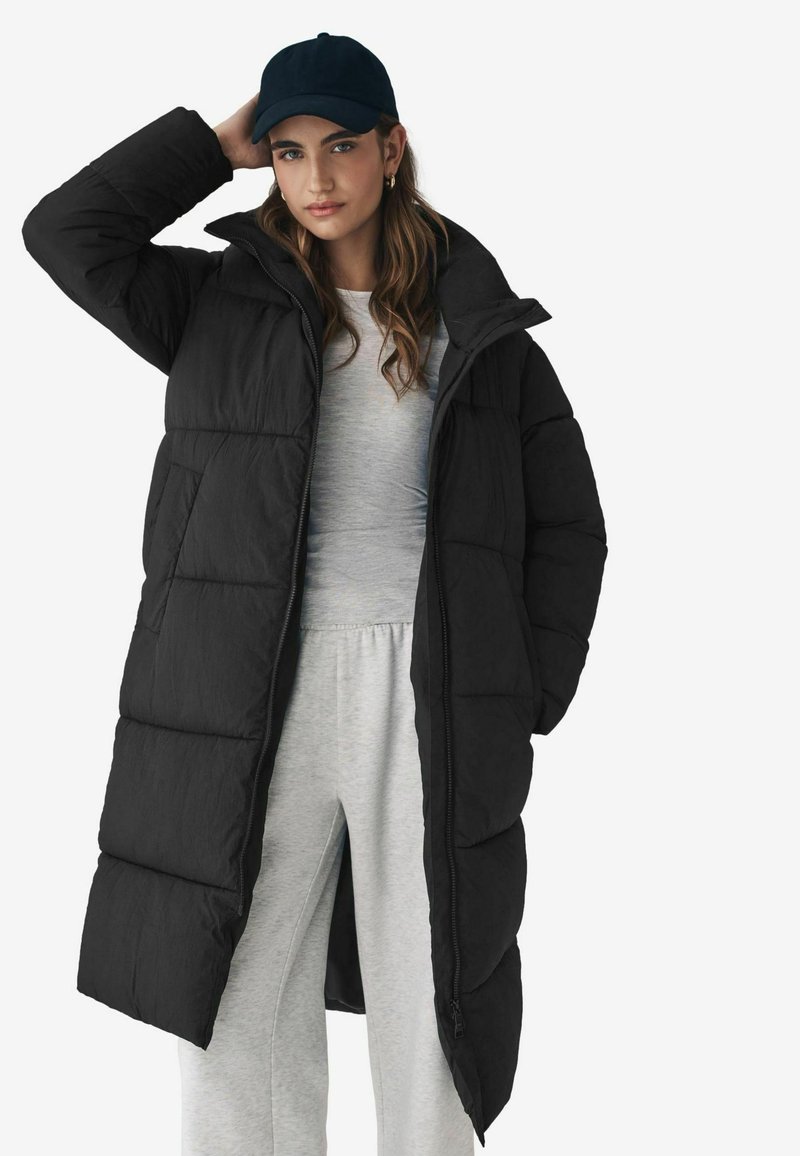 Cappotto piumino nero con un design lungo e texture trapuntata, dotato di un colletto alto e chiusura con zip. Abbinato a un top grigio e pantaloni della tuta.