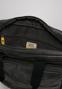 Schwarze Ledertasche mit strukturierter Oberfläche, featuring einer Innentasche mit gelben Akzenten und einem Reißverschluss. Innenfutter mit sichtbarem Markenlabel.