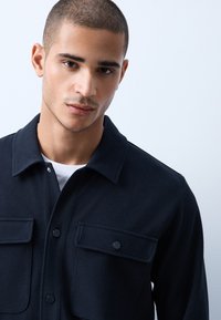 Jeune homme aux cheveux courts portant une chemise noire à boutons avec deux poches poitrine, debout devant un fond clair uni.