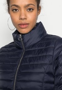 Giacca puffer trapuntata blu navy con colletto alto, chiusura con zip e cuciture orizzontali. Tessuto liscio e lucido con dettaglio della zip metallica.