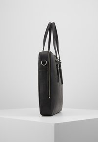 Bolso tote de cuero negro con una superficie texturizada, cremallera plateada y asas dobles, mostrado de lado sobre un pedestal blanco.