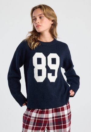 Femme portant un pull marine avec le numéro "89" en blanc et un pantalon à carreaux rouges, les mains dans les poches, regardant légèrement sur le côté sur un fond uni.