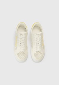 Fehér sportcipők geometriai kivágásos mintákkal, fehér fűzőkkel és sárga díszítéssel a sarokrészen, felülről nézve, fehér háttéren.