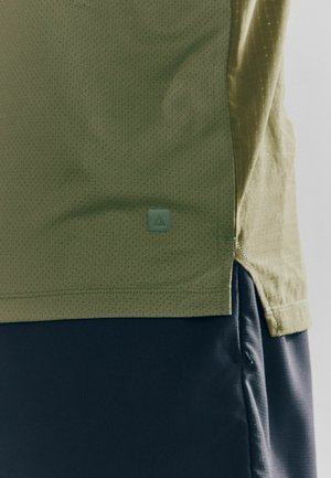 Chemise olive verte à manches courtes en tissu texturé et respirant. Elle présente un petit badge logo vert et une fente sur le côté pour faciliter les mouvements.