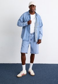 Stribet denimskjorte og matchende shorts i lyseblå. Hvid tank top med palmelogo under. Beiges kasket og cremefarvede sneakers med røde accenter.