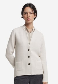 Jasno beżowy sweter narzutowy z teksturowanym wzorem, z dekoltem w serek, dwoma przednimi kieszeniami i trzema dużymi guzikami.