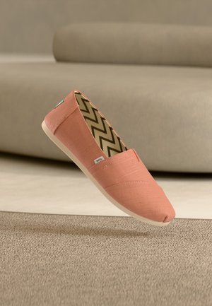Koralfarvet slip-on sko i lærred med zigzag-mønstret indersål, svævende over beige tæppe foran en taupefarvet sofa.