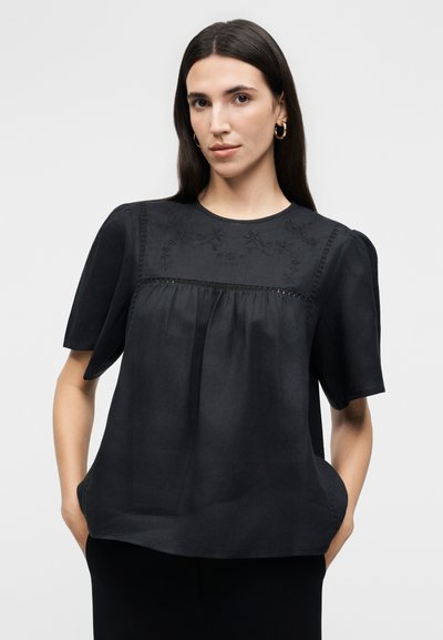 Γυναίκα με μακριά σκούρα μαλλιά φοράει μαύρο μπλουζάκι με κοντά μανίκια και κεντήματα με λουλούδια στο στήθος, στέκεται με τα χέρια στις τσέπες μπροστά σε φωτεινό φόντο.