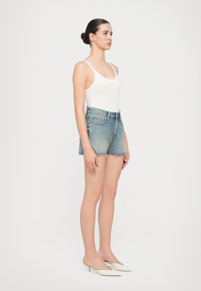 Hvit tanktopp, denimshorts med slitt kant, og hvite slip-on sko, står mot en ensfarget bakgrunn. Shortsene har en falmet vask.