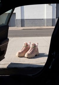 Coppia di stivali da trekking beige con lacci verdi sul marciapiede fuori dalla portiera aperta di un'auto vicino a una strada e a un edificio bianco.